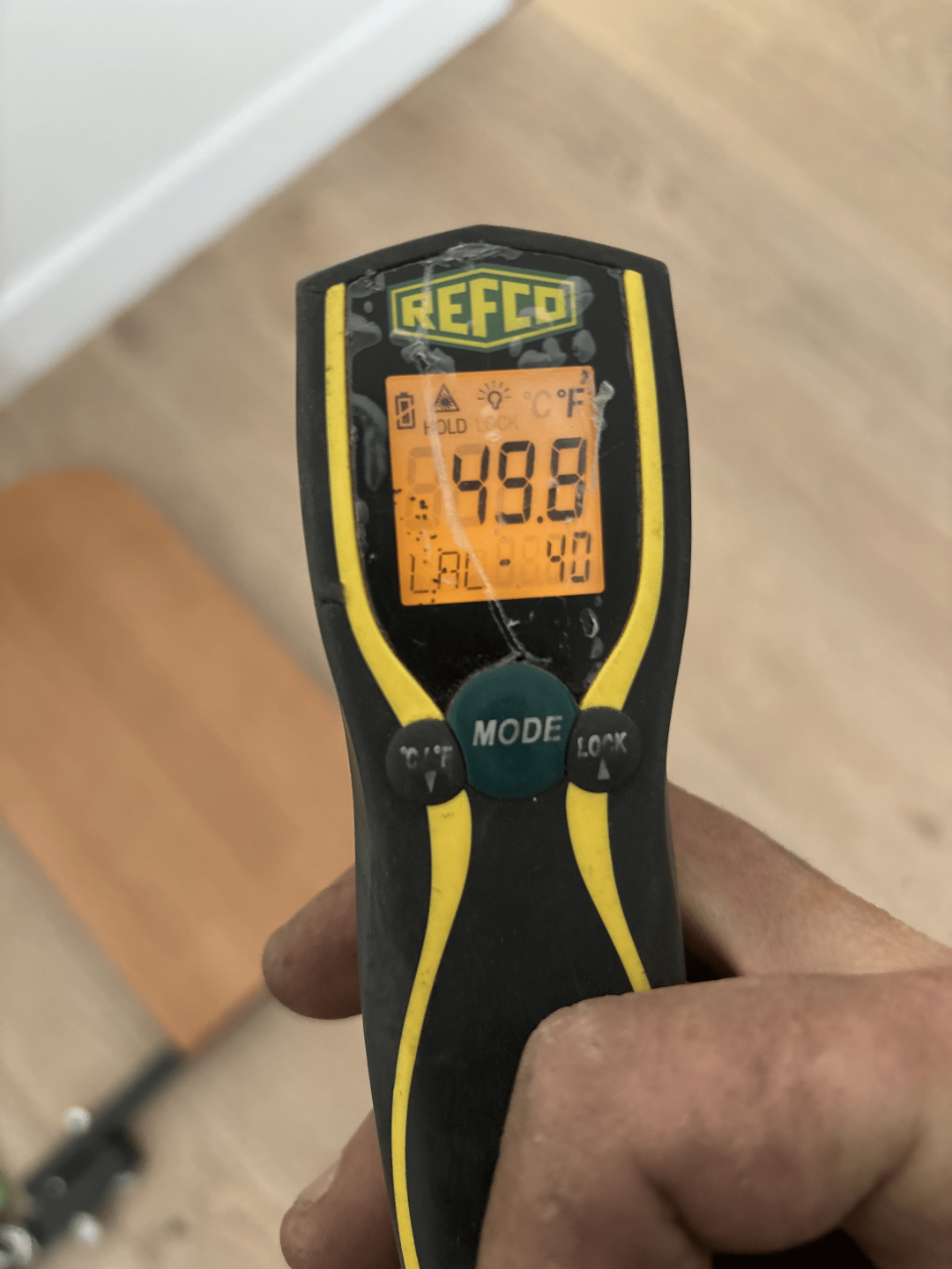 A person holds a digital thermometer above a wooden floor, indicating a focus on temperature measurement.